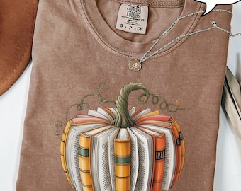 Könnte beinhalten: Espressofarbenes T-Shirt mit einem Kürbisdesign aus Büchern. Der Kürbis hat einen grünen Stiel und orange, gelbe und weiße Buchseiten. Eine silberne Halskette mit einem kleinen Anhänger ist auf dem T-Shirt drapiert.
