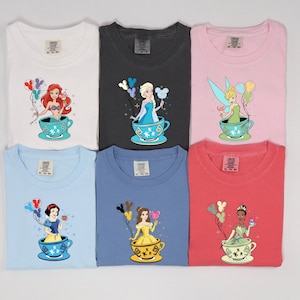 May include: Six different t-shirts featuring Disney princesses in teacups with balloons. The princesses are Ariel, Elsa, Tinkerbell, Snow White, Belle, and Tiana. The shirts are in various colors, including white, black, pink, light blue, blue, and red.