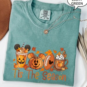 May include: Light green t-shirt with a fall-themed graphic. The design includes a pumpkin spice latte, Mickey Mouse pumpkins, a football, and a mug of hot cocoa. The text "Tis The Season" is printed in orange.