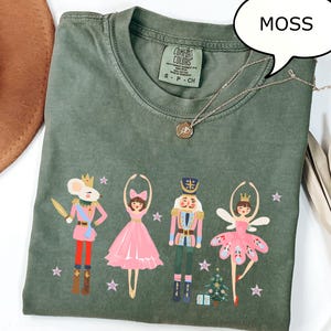 Op de afbeelding: Een mosgroen T-shirt met een Notenkraker-thema ontwerp. De afbeelding bevat illustraties van een muizenkoning, ballerina, notenkraker en een andere ballerina, samen met een kleine kerstboom en sterren. Het woord "MOSS" staat in een tekstballon.