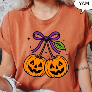 May include: Orange t-shirt featuring a Halloween-themed design. The graphic shows two orange jack-o'-lanterns with smiling faces, connected by stems and a purple bow. A green leaf and stars complete the design. The word "YAM" is in a speech bubble.