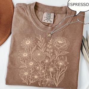 Op de afbeelding: Espresso-kleurig T-shirt met een bloemenontwerp. Het shirt heeft een ronde hals en korte mouwen. Een zilveren ketting met een hanger ligt op het shirt. Het shirt is gelabeld met "Comfort Colors". Het woord "Espresso" staat in een tekstballon.