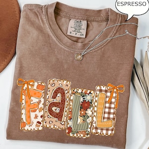 May include: Espresso-coloured t-shirt featuring the word "FALL" in a decorative design. The letters are filled with autumn-themed patterns, including pumpkins, floral prints, and plaid. A silver necklace with a pendant is draped across the shirt.