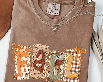May include: Espresso-colored t-shirt with the word "FALL" in a decorative design. The letters are filled with fall-themed patterns, including pumpkins, floral prints, and plaid. A silver necklace with a pendant is draped across the shirt.