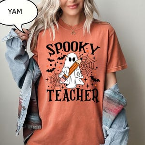 May include: An orange short-sleeve t-shirt featuring a graphic of a white ghost holding a pencil, surrounded by black bats, spider webs, and stars. The text SPOOKY TEACHER is printed in black above and below the ghost. A denim jacket is worn over it.