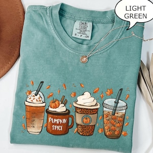 May include: Light green t-shirt featuring a graphic of four autumn-themed drinks, including a pumpkin spice latte. The shirt has a crew neck and short sleeves. A silver necklace with a small pendant is on the shirt.