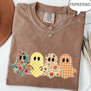 May include: Espresso-colored Comfort Colors t-shirt with four ghost appliqués. Each ghost has a different floral or gingham pattern. The shirt has a crew neck and short sleeves. A silver necklace with a small pendant is draped across the shirt.