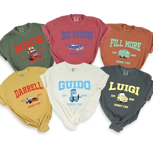 Puede incluir: Seis camisetas de diferentes colores con personajes y nombres de Cars. Las camisetas est&aacute;n todas dobladas y dispuestas sobre un fondo blanco. Los personajes incluyen Mack, Doc Hudson, Fill More, Darrell, Guido y Luigi.
