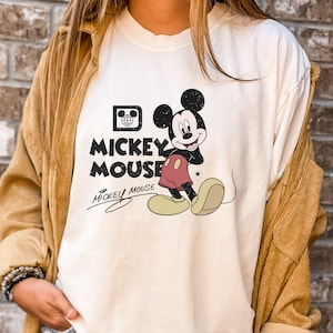 May include: White t-shirt with a vintage Mickey Mouse graphic. The graphic features Mickey Mouse in a red and black outfit with a yellow star on his shorts. The text "Mickey Mouse" is printed in black letters below the graphic.
