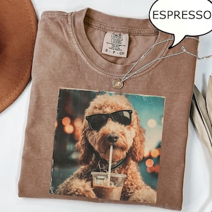 Puede incluir: Camiseta color espresso con un gráfico de un perro con gafas de sol bebiendo de una taza transparente. La camiseta tiene cuello redondo y mangas cortas. Un collar plateado con un pequeño colgante está sobre la camiseta. La palabra "ESPRESSO" está en una burbuja.
