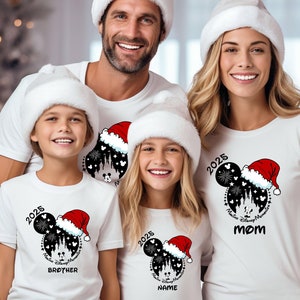 May include: Family matching Christmas shirts featuring a Mickey Mouse design with fireworks, Cinderella Castle, and a Santa hat.  The shirts are white with black and red accents.  The year '2025' is printed on the adult shirts, while the children's shirts display '2026' and '2025' respectively.  The text 'MOM', 'BROTHER', and 'NAME' is also included on the shirts.