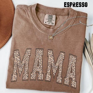 May include: A brown t-shirt with the word "MAMA" printed in a leopard print pattern. The shirt is made of a soft, comfortable material and has a relaxed fit.
