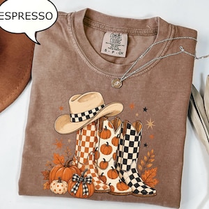 Könnte beinhalten: Ein espressofarbenes T-Shirt mit einem Grafikdruck, der einen Cowboyhut und drei Cowboystiefel mit Karomustern und Kürbissen zeigt. Das T-Shirt hat einen Rundhalsausschnitt und ein Comfort Colors Markenetikett am inneren Kragen.