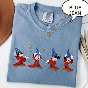 May include: Blue jean colored t-shirt featuring four Mickey Mouse characters dressed as wizards. Each character wears a blue wizard hat and a red robe. The shirt has a Comfort Colors tag and a gold necklace.