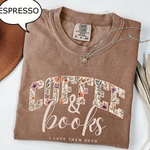 May include: A brown short-sleeve t-shirt with a crew neckline. The shirt features the text COFFEE & books I LOVE THEM BOTH in a floral pattern. A small silver necklace with a circular pendant rests on the collar area of the garment.