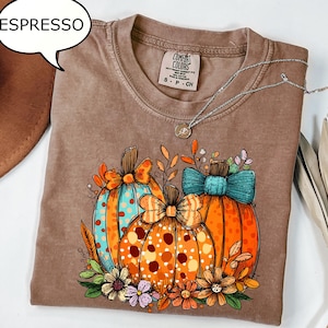 Könnte beinhalten: Ein braunes Kurzarm-T-Shirt mit einem Grafikdruck von drei Kürbissen mit Schleifen, umgeben von Herbstblättern und Blumen. Das T-Shirt hat ein Comfort Colors-Etikett am Halsausschnitt und eine Sprechblase mit dem Text ESPRESSO.