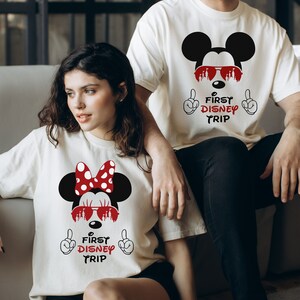 May include: White t-shirts with a Mickey Mouse and Minnie Mouse graphic. The graphic features Mickey Mouse wearing sunglasses and Minnie Mouse wearing a red bow and sunglasses. The text on the shirts reads "First Disney Trip".