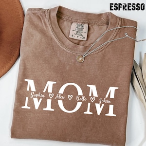 May include: Espresso-colored t-shirt with the word "MOM" in white block letters. Below the word "MOM" are the names Sophia, Alex, Belle, and Johan, each with a heart symbol. A silver necklace with a pendant is draped on the shirt.
