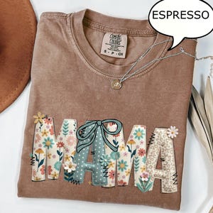 以下が含まれることがあります： エスプレッソ色のTシャツに、花柄と模様の生地で「MAMA」の文字がアップリケされています。Tシャツはクルーネックで半袖です。小さなペンダントが付いたシルバーのネックレスがTシャツの上にかけられています。