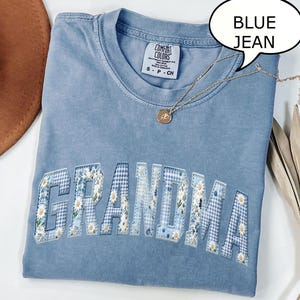 May include: A blue jean colored t-shirt with the word "GRANDMA" in a floral and gingham patchwork design. The shirt has a crew neck and short sleeves. A gold necklace with a pendant is draped across the shirt.
