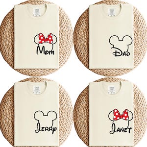 May include: Four cream-colored t-shirts with black Mickey Mouse head outlines and names. One shirt has a red and white polka dot bow with the word "Mom". Another has "Dad". The third has "Jerry", and the fourth has a bow and the name "Janet".