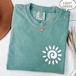 May include: A light green t-shirt with a white sun graphic. The t-shirt has a crew neck and short sleeves. The sun graphic has a spiral in the center.