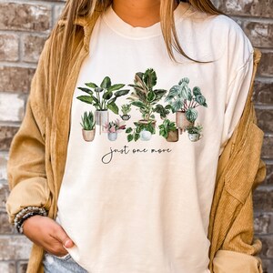May include: A white t-shirt with a watercolor illustration of potted plants and the text "just one more".