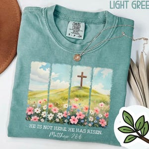 May include: A light green t-shirt featuring a graphic of a cross on a hill with flowers and the text "HE IS NOT HERE. HE HAS RISEN. Matthew 28:6".