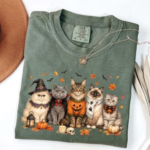 Puede incluir: Una camiseta verde salvia con un gráfico de cinco gatos vestidos con disfraces de Halloween, incluyendo un sombrero de bruja, una capa de vampiro, un suéter de calabaza, una bufanda de fantasma y una pajarita, rodeados de hojas, murciélagos y calavera.