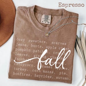 Könnte beinhalten: Ein espressofarbenes Kurzarm-T-Shirt mit Rundhalsausschnitt. Das Shirt ist mit weißem Text bedruckt, der herbstliche Begriffe wie cozy sweaters, scarves, jeans, boots, apple cider, pumpkin patches, leaves, football und bonfires auflistet.