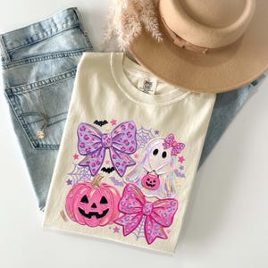 May include: A cream-colored t-shirt featuring a graphic print of a pink jack-o'-lantern, a white ghost holding a small pumpkin bucket, and two large purple and pink leopard-print bows, accented with spiderwebs, bats, and small stars.