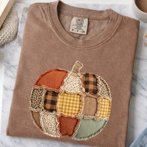May include: A brown short-sleeve t-shirt featuring a Comfort Colors label at the neck. The shirt displays a large, three-dimensional pumpkin applique made from various fabric patches, including plaid, leopard print, floral, and solid textures.