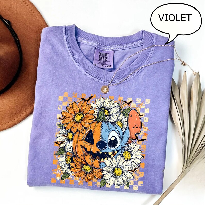 May include: A violet Comfort Colors t-shirt featuring a Halloween-themed graphic. The design includes a jack-o'-lantern, a blue cartoon character, orange and white flowers, and bats. A gold necklace is draped across the shirt.