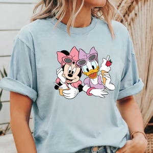 Puede incluir: Una camiseta azul claro con una imagen de dibujos animados de Minnie Mouse y Daisy Duck con gafas de sol y sosteniendo conos de helado.