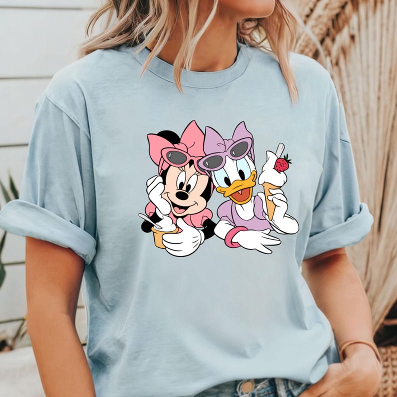 Puede incluir: Una camiseta azul claro con una imagen de dibujos animados de Minnie Mouse y Daisy Duck con gafas de sol y sosteniendo conos de helado.