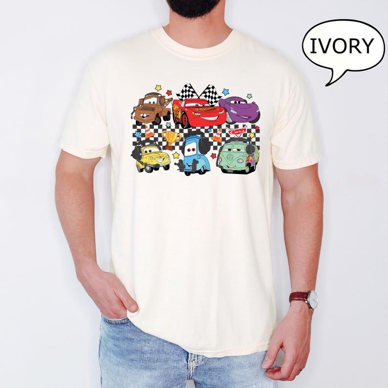 May include: A white t-shirt with a checkered pattern and a graphic of cartoon cars. The cars are red, blue, green, purple, and brown. The text "Cars" is on the shirt.