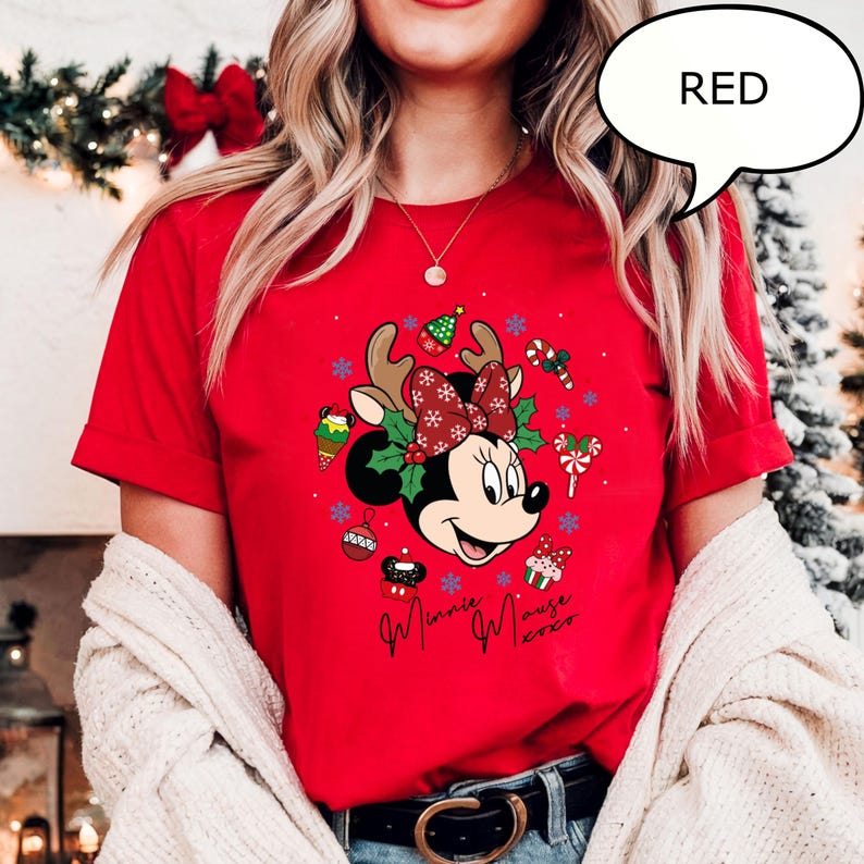 May include: A red t-shirt featuring a festive Minnie Mouse design. Minnie Mouse is wearing a reindeer antler headband and a bow. The shirt has the text "Minnie Mouse 2020" in cursive. The word "RED" is in a speech bubble.
