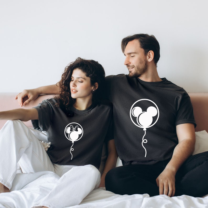 Pode incluir: Um casal vestindo camisetas pretas combinando com desenhos de bal&otilde;es do Mickey Mouse e da Minnie Mouse brancos. A mulher est&aacute; usando um la&ccedil;o em seu bal&atilde;o e o homem est&aacute; usando orelhas do Mickey Mouse em seu bal&atilde;o.