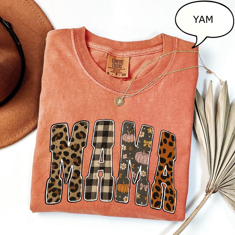 May include: Coral-colored Comfort Colors t-shirt with the word "MAMA" in a mix of leopard print, plaid, and floral patterns. A gold necklace with a pendant rests on the shirt. The shirt is a small size.
