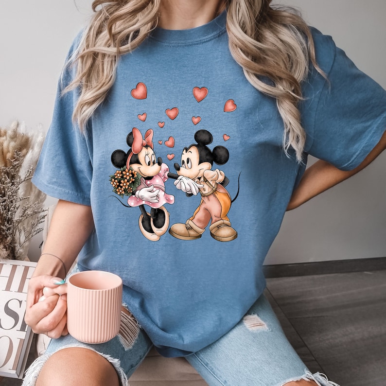 May include: A blue t-shirt with a graphic of Mickey Mouse and Minnie Mouse holding hands.  There are red hearts scattered around them.