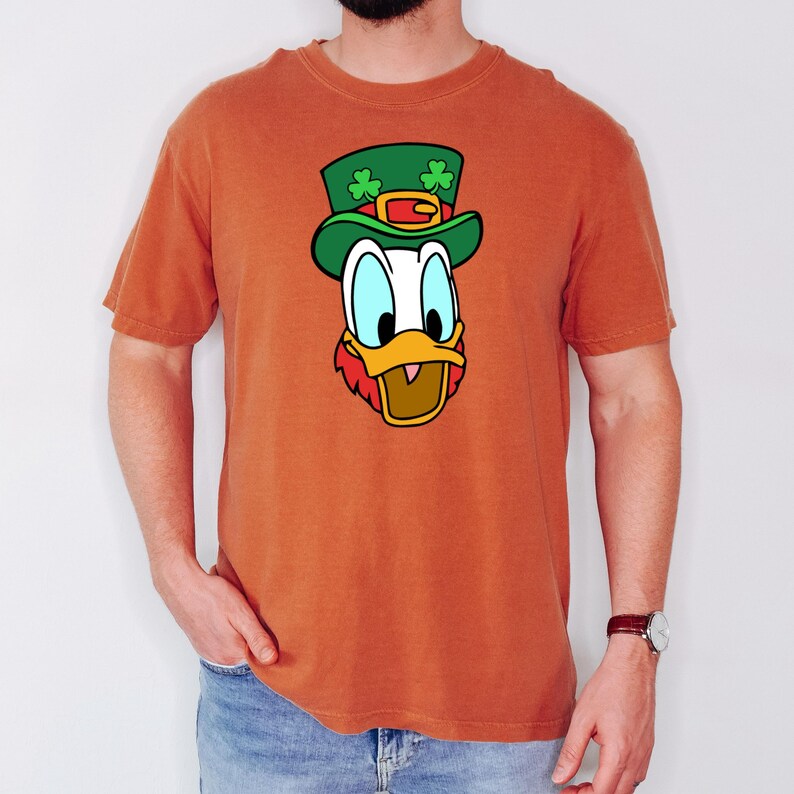 Puede incluir: Camiseta naranja con un dibujo de un pato de dibujos animados con un sombrero de duende verde con tr&eacute;boles. El pato tiene un pico amarillo, barba roja y ojos azules. La camiseta es de manga corta y tiene cuello redondo.