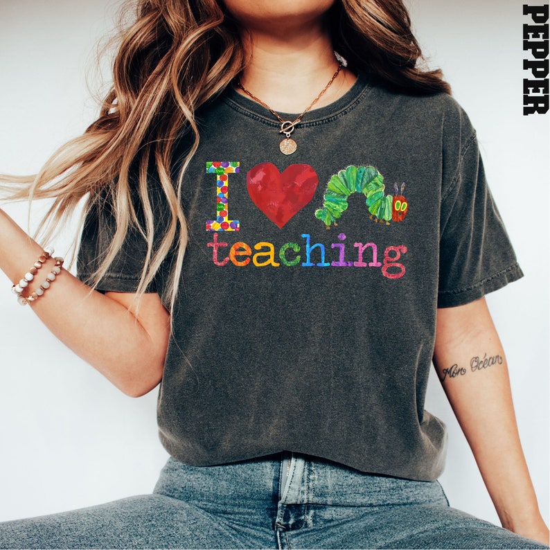 Op de afbeelding: Een donkergrijs T-shirt met de tekst "Ik hou van lesgeven" in regenboogkleuren. Naast de tekst bevinden zich een rood hart en een groene rups met een rood gezicht. Het woord "Pepper" staat in de rechterbovenhoek van de afbeelding.