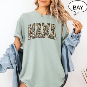 May include: A light green t-shirt with a camouflage print of the word "MAMA" in a collegiate font.