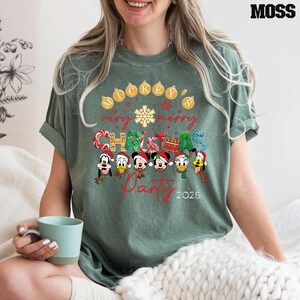 K&ouml;nnte beinhalten: Ein gr&uuml;nes T-Shirt mit einem festlichen Mickey Mouse Weihnachtsparty-Design. Der Text lautet "Mickey's Very Merry Christmas Party 2025" und zeigt Mickey Mouse, Minnie Mouse, Donald Duck, Goofy und Pluto.