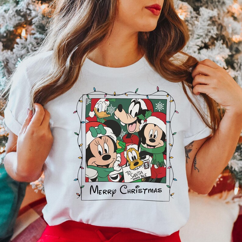 May include: White t-shirt with a Christmas-themed graphic featuring Mickey Mouse, Minnie Mouse, Donald Duck, Goofy, and Pluto. The graphic is a Polaroid-style photo with a string of Christmas lights around it. The text "Merry Christmas" is printed below the graphic.