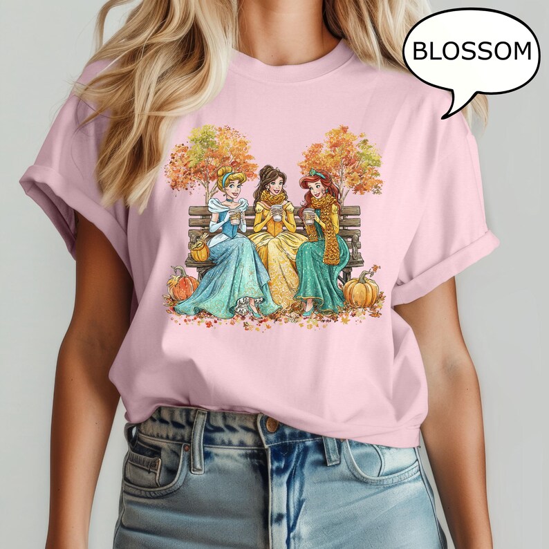 May include: Pink t-shirt featuring a fall-themed illustration of three princesses sitting on a bench, holding cups, with pumpkins and autumn leaves. The word "BLOSSOM" is in a speech bubble.