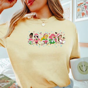 Puede incluir: Una camiseta amarilla con un gr&aacute;fico navide&ntilde;o que presenta a seis princesas Disney con gorros de Pap&aacute; Noel y sosteniendo regalos. Las princesas est&aacute;n rodeadas de luces navide&ntilde;as y copos de nieve. El texto "Jingle" aparece dos veces en la camiseta.