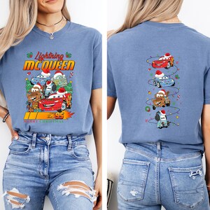 May include: Blue t-shirt with a festive graphic of Lightning McQueen and other Cars characters. The front displays "Lightning McQueen" and "Merry Christmas". The back of the shirt shows the characters wearing Santa hats and Christmas lights.