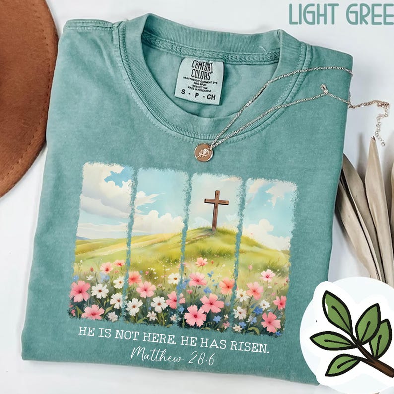 May include: A light green t-shirt featuring a graphic of a cross on a hill with flowers and the text "HE IS NOT HERE. HE HAS RISEN. Matthew 28:6".
