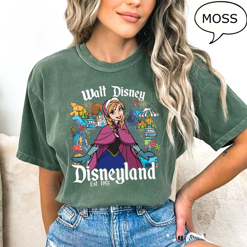 May include: Moss green t-shirt with a graphic of Anna from Disney's Frozen, and the words "Walt Disney Disneyland Est. 1955". The design includes park attractions and a bear. The shirt is a moss green colour.